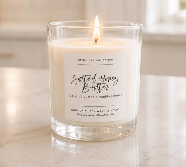 A lit candle in a clear glass jar with a white label reading 'little love collective Salted Honey Butter' on a light marble surface in a softly blurred indoor setting.